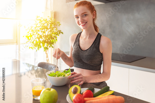 Athletic young red haired woman in the home kitchen eating a healthy salad photo