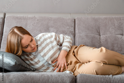 Young woman has abdominal pain lying on couch in working day in office. Acute pain in bloating pms. Teenage girl with pain problems bowel disease