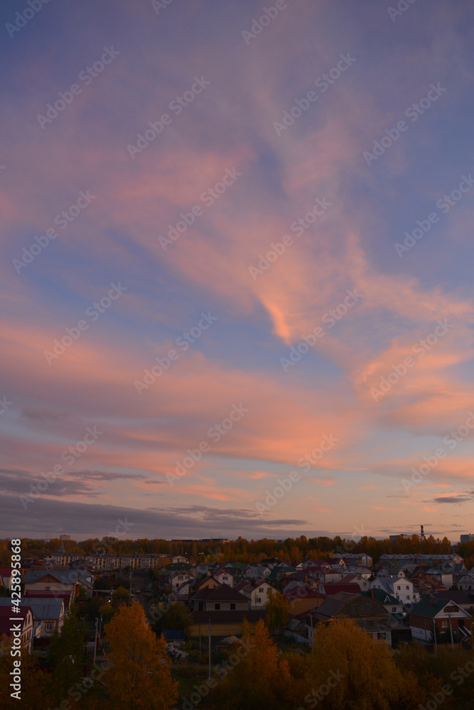 Beautiful landscape with town in autumn evening. Pink clouds in blue sky, houses and trees with golden foliage