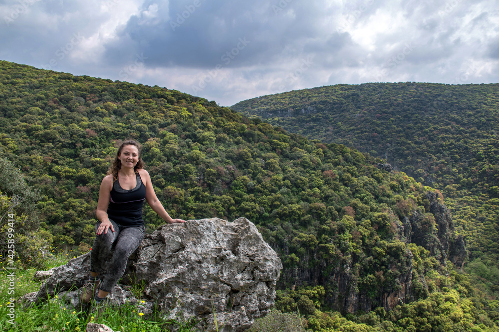 woman traveler sitting on rock and looking at amazing green hills and forest, hiking trip through Israel mountains, wanderlust travel concept, space for text, atmospheric epic moment