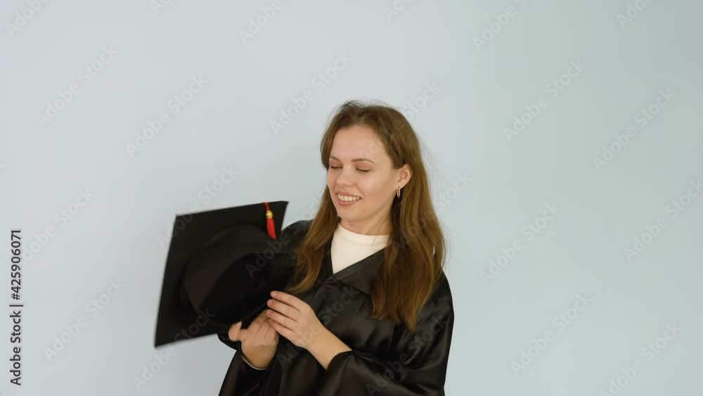 A young caucasian female student in a black robe jokingly plays with a square hat and tickles her nose with a brush. White background A young caucasian female student in a black robe jokingly plays with a square hat and tickles her nose with a brush. White background