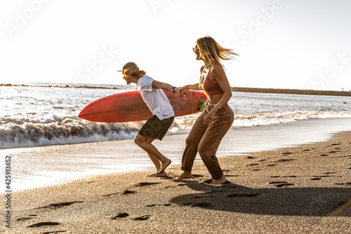 Beautiful happy surfer couple having fun on the sandy beach, running with surfboard to the waves.