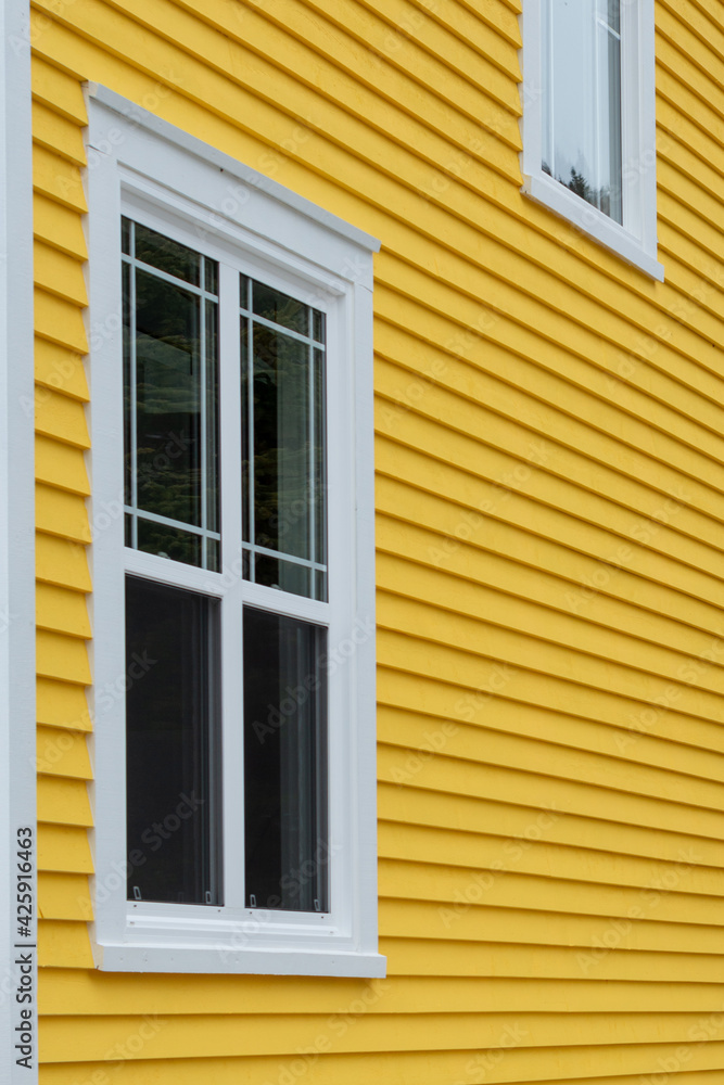 The exterior of a bright yellow narrow wooden horizontal clapboard wall of a house with two vinyl windows. The trim on the glass panes is white in colour. The outside boards are textured pine wood. The exterior of a bright yellow narrow wooden horizontal clapboard wall of a house with two vinyl windows. The trim on the glass panes is white in colour. The outside boards are textured pine wood.