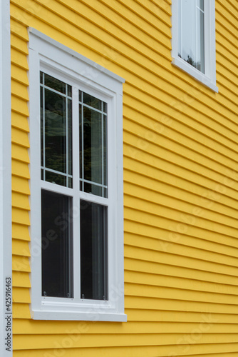 The exterior of a bright yellow narrow wooden horizontal clapboard wall of a house with two vinyl windows. The trim on the glass panes is white in colour. The outside boards are textured pine wood.  