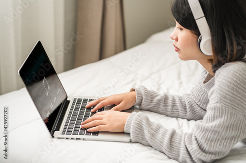 young woman in casual wear lying listening to music on the laptop with a white headphone in the bedroom fun, female lifestyle in holiday.
