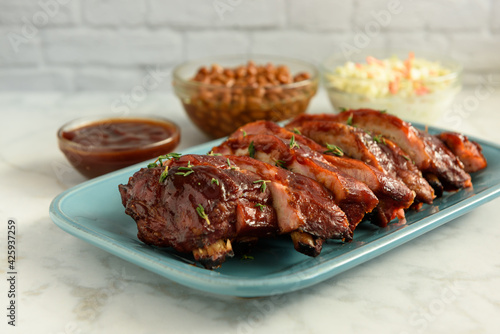 Close up of sliced smoked pork BBQ ribs on a blue plate with sides of BBQ sauce, baked beans and coleslaw on a white marble table and subway tile background