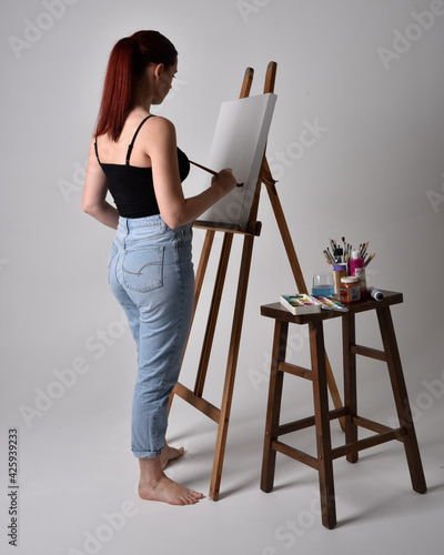 Full length portrait of a red haired artist girl wearing casual jeans and tank top. standing pose painting a canvas on an easel, against a studio background.