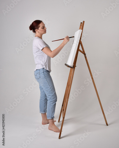 Full length portrait of a red haired artist girl wearing casual jeans and white shirt. standing pose painting a canvas on an easel, against a studio background.