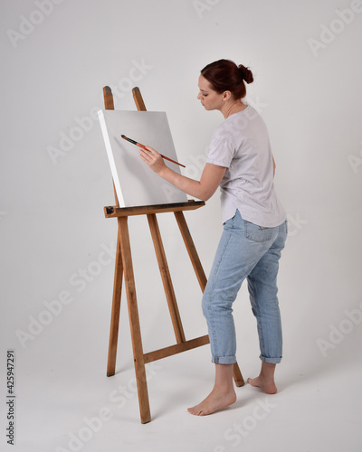 Full length portrait of a red haired artist girl wearing casual jeans and white shirt. standing pose painting a canvas on an easel, against a studio background.