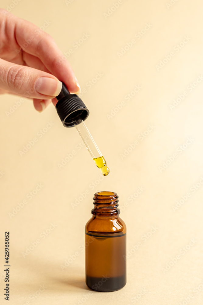 female hand with Dropper of essential oil, aromatherapy essence, beuty serum or medicinal liquid on beige background. Unbranded bottles for your design. female hand with Dropper of essential oil, aromatherapy essence, beuty serum or medicinal liquid on beige background. Unbranded bottles for your design.