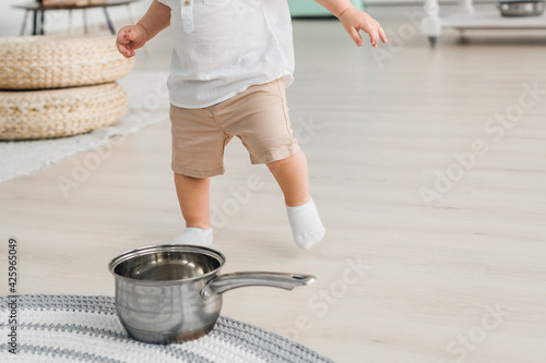 The baby boy runs to the pan, which stands on the floor. The child plays with kitchen utensils