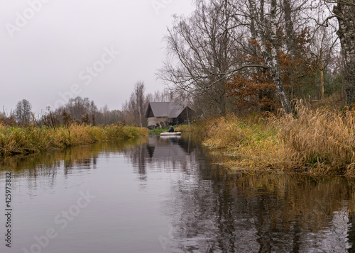 gray and cloudy day, fisherman in a white boat, river bank with bare trees and bushes, shore reflection in river water
