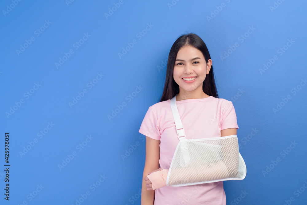 Portrait of young woman with an injured arm in a sling over blue background in studio, insurance and healthcare concept Portrait of young woman with an injured arm in a sling over blue background in studio, insurance and healthcare concept