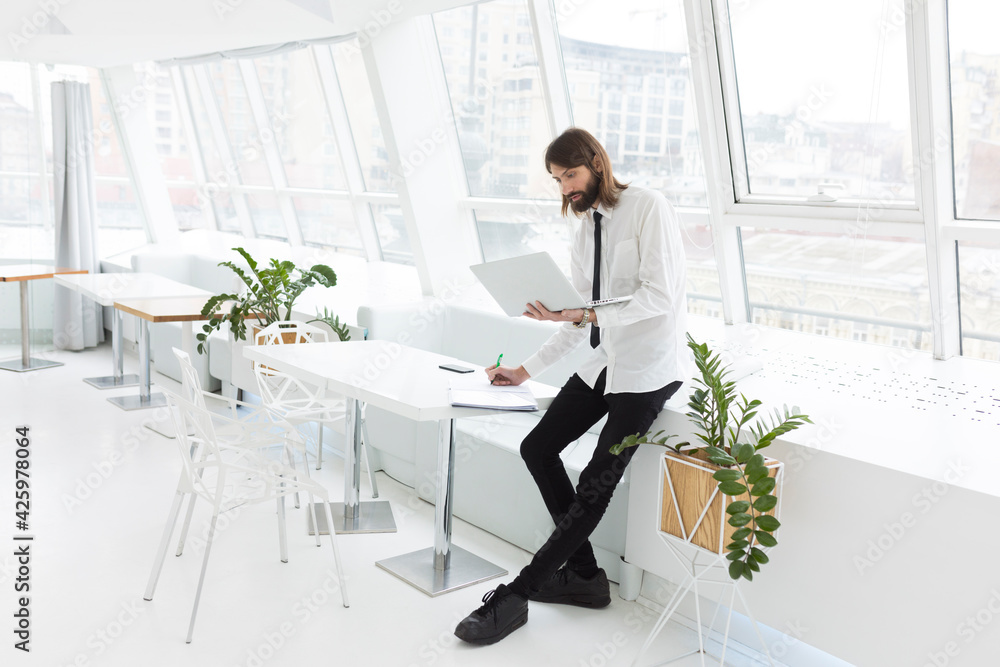 business man in a white shirt and tie stands working with documents and laptop in a modern office by the window
business man in a white shirt and tie stands working with documents and laptop in a modern office by the window