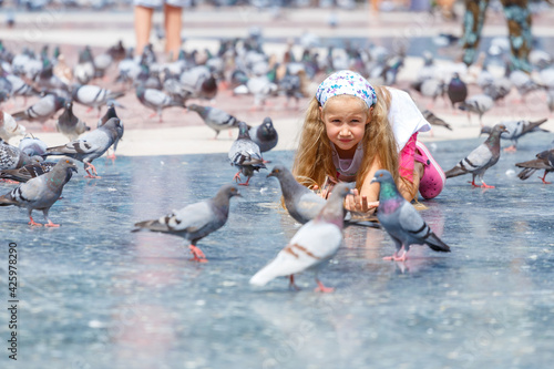 A beautiful Caucasian blonde girl with hair, 6-7 years old, feeding pigeons at Plaza de Catalunya in Barcelona. Bird watching. Summer vacation, travel to Europe. Childhood, carelessness. Real life. photo