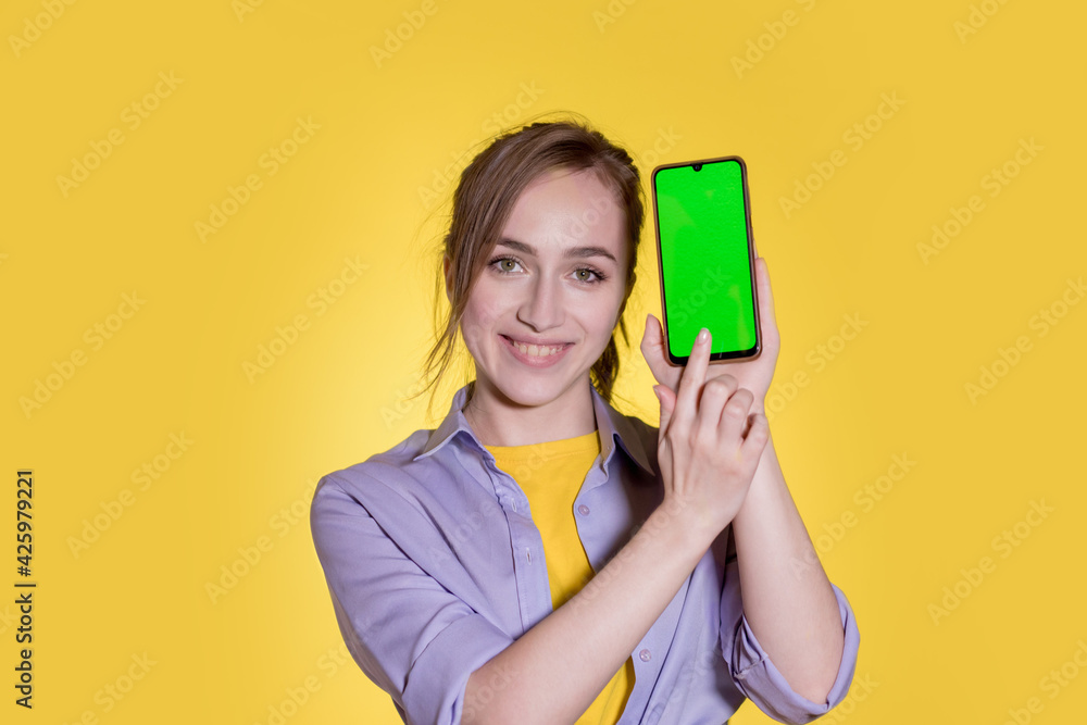 Young woman pointing at green screen on smartphone on yellow background Young woman pointing at green screen on smartphone on yellow background