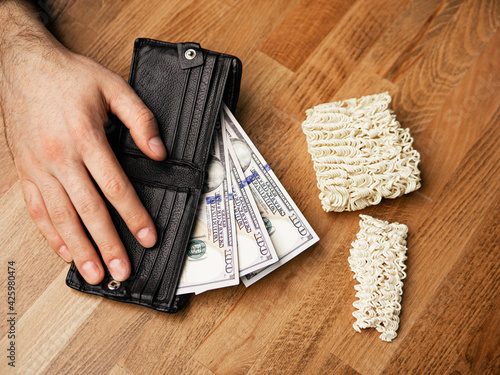 Young male hand is on the black wallet with dollar currency. Instant noodle (ramen) in pocket as poverty concept. Financial problems, crisis and bankruptcy. Coronavirus and society living issues.