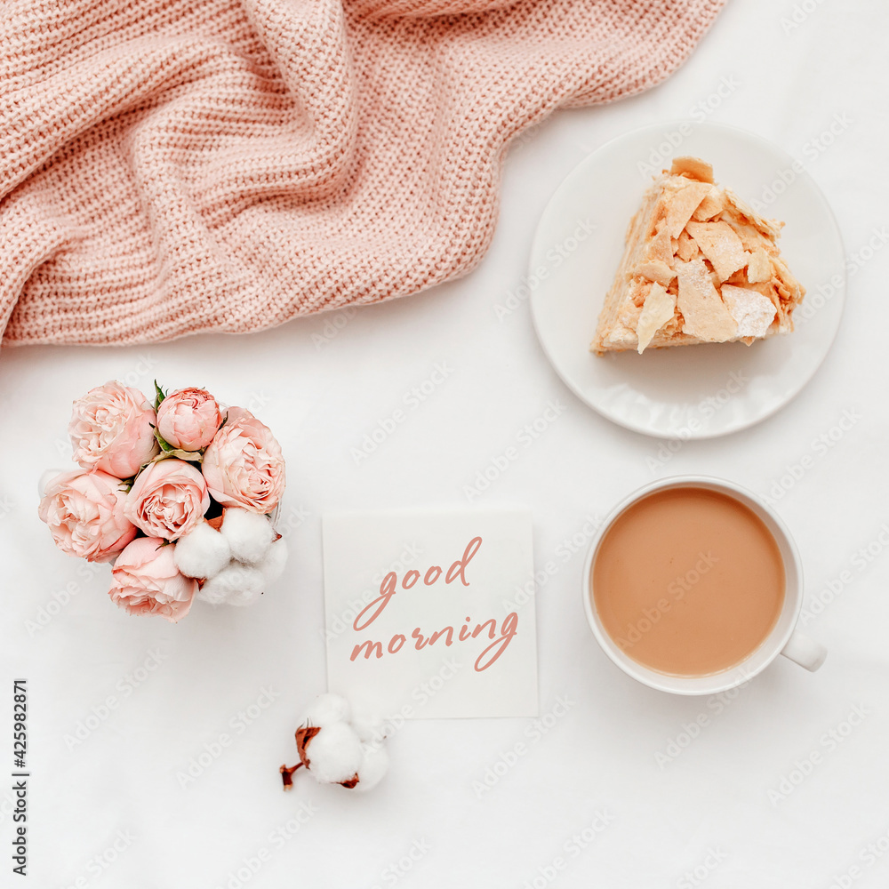 Cup of coffee with milk, piece of cake, rose flowers, pink knitted plaid or blanket, card with text GOOD MORNING. Breakfast in bed. Stylish home interior decor. Flat lay, top view. Cup of coffee with milk, piece of cake, rose flowers, pink knitted plaid or blanket, card with text GOOD MORNING. Breakfast in bed. Stylish home interior decor. Flat lay, top view.