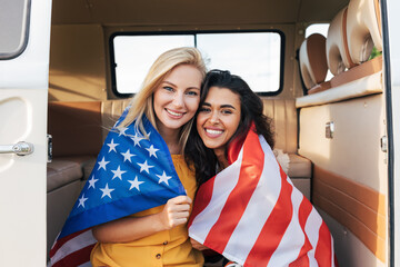 Two young female friends with American flag sitting in a camper van. Beautiful women with USA flag looking at camera.