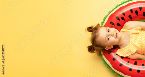 Top view of funny little child girl dressed in a swimsuit lying on an inflatable color red ring on yellow studio background. Summer travel concept. Banner, mock up, copy space