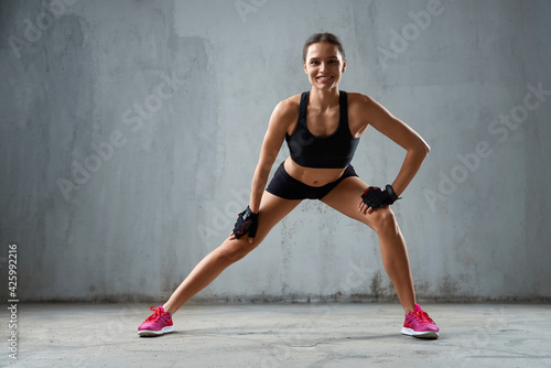 Front view of young female wearing sports underwear, training and smiling. Muscular sportswoman with perfect body practicing legs stretching and looking at camera in empty hall, gray loft interior.