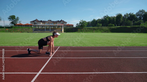 Attractive fit female sprinter on racetrack at stadium starting running race. Side view of active athletic woman in sprinter position preparing for run in summer sunny day. Concept of sport.