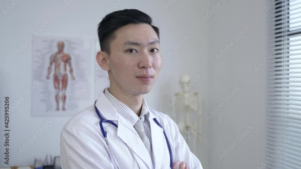 shoulder shot handsome asian physician is smiling at the camera with confidence by the window at a bright workplace. shoulder shot handsome asian physician is smiling at the camera with confidence by the window at a bright workplace.