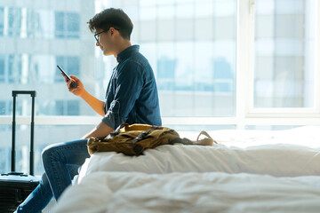 young adult asian traveller male casual cloth with jean jacket hand use smartphone conversation on white soft bed with luggage bagpack on vacation weekend travel concept