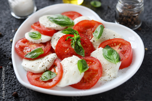 Fresh and delicious caprese salad with sliced tomatoes, mozzarella cheese, basil leaves and olive oil on a white heart-shaped plate. Selective focus