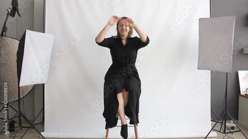 photo studio with white background. girl in black dress is sitting on the high chair and smiling cute. funny conversation with model before shooting. making off interview. lifestyle concept photo