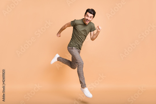 Full size profile photo of impressed brunet guy jump run wear t-shirt pants sneakers isolated on pastel beige color background