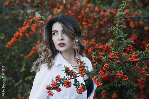 Sensual young woman in white shirt with makeup and curly hair posing near bush with orange berries in the park.