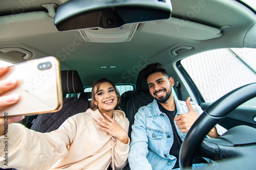 road trip, leisure, couple, technology and people concept - happy indian man and woman driving in cabriolet car and taking picture with smartphone on selfie stick