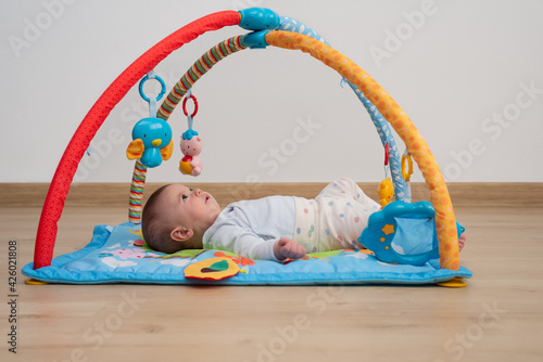 Cute 5 months baby girl lying face up on a blue play mat while is looking at toys of a baby gym.   Games for psychomotor development.  photo