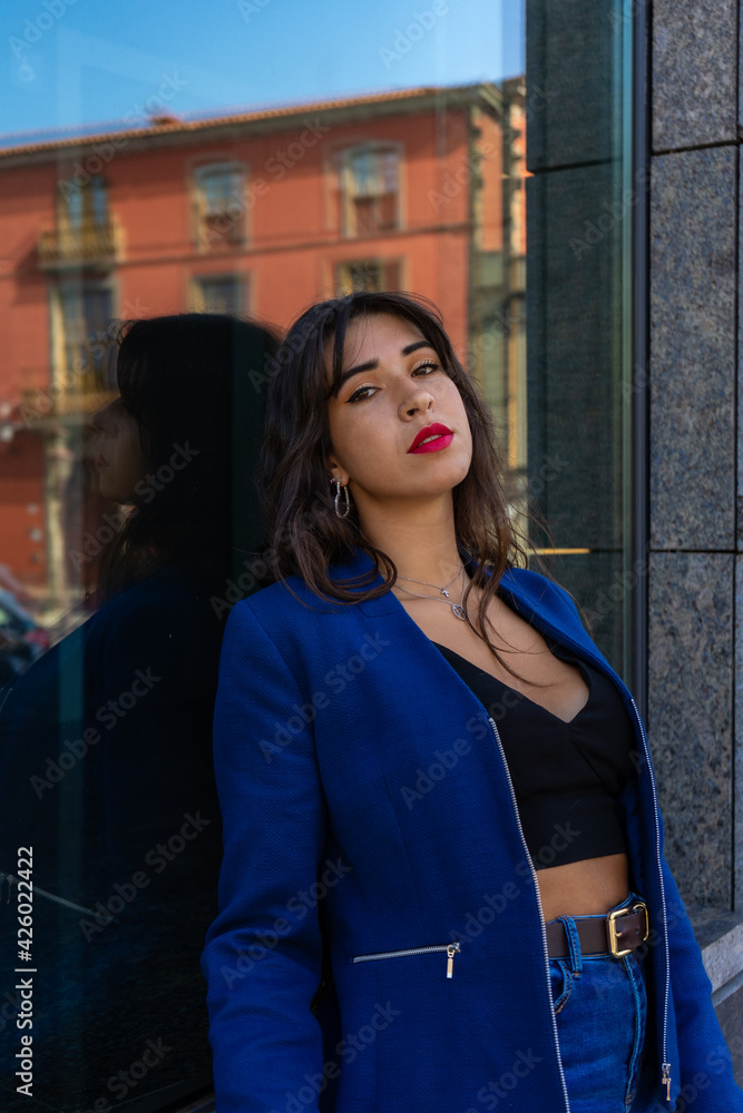 Fototapeta premium Young woman posing for a portrait in the background looks a glass. She wears an American jacket and jeans. The photo was taken in La Laguna, Tenerife.