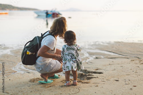 Beautiful mother and baby outdoors Mum and her child together enjoying seaview. Back view. Woman with backpack and cute girl in beautiful dress. Travel with toddlers, family day concept