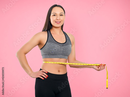 A slender woman measures her waist with a measuring tape. n a pink background.