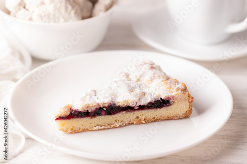 A piece of handmade grated cherry pie on a white plate. Sprinkled on top with powdered sugar. White wood background. Close-up. In the background a white cup and white cookies in a bowl 