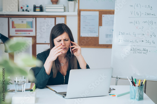 Upset female professor talking on mobile phone on break with online class. Tired teacher is working from home, complaining on phone, angry and displeased with teleconferencing and e-learning students photo