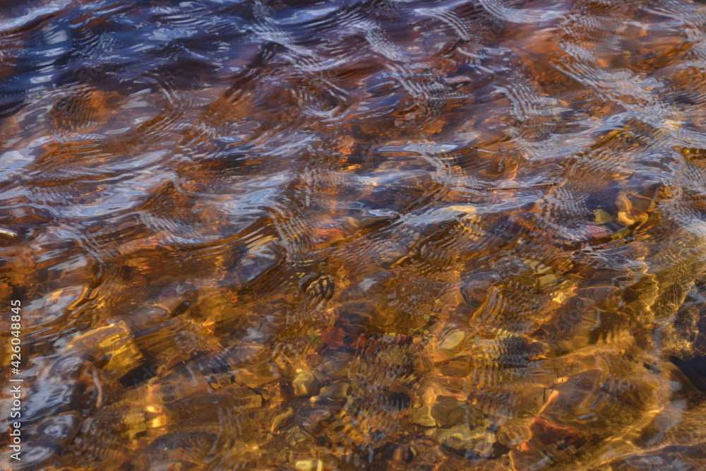 water texture pattern in lake in spring sunlight. Through the clear water you can see small stone pebbles on the shore. small waves in the water. water texture pattern in lake in spring sunlight. Through the clear water you can see small stone pebbles on the shore. small waves in the water.