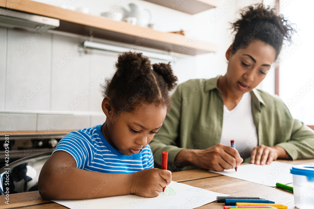 Black mid woman drawing with her focused daughter at home kitchen Black mid woman drawing with her focused daughter at home kitchen