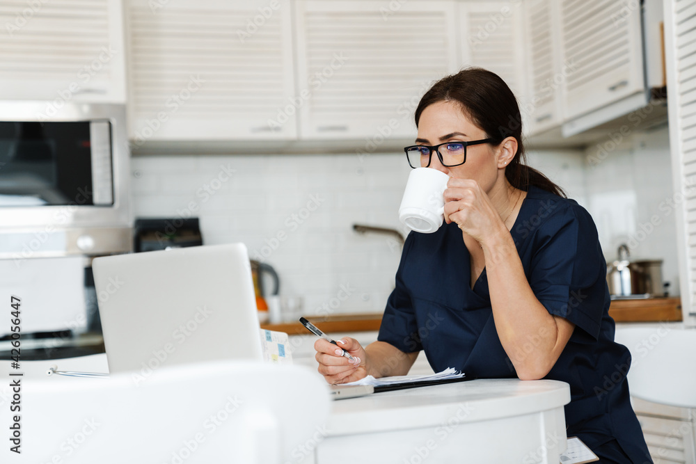 The thoughtfully woman in a medical suit sitting in front of a laptop and drinking something in the kitchen The thoughtfully woman in a medical suit sitting in front of a laptop and drinking something in the kitchen