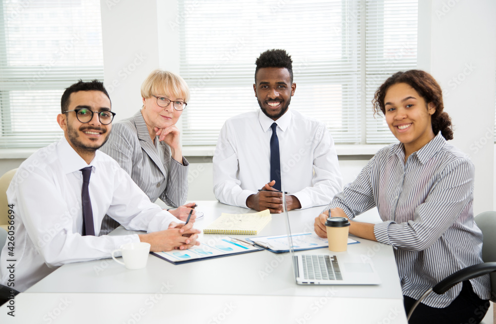 Multy-ethnic group of business people working while sitting at the office desk and smiling at camera Multy-ethnic group of business people working while sitting at the office desk and smiling at camera