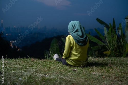 Pretty girl sitting on grass, relaxing, enjoying Kuala Lumpur city view during sunset. Portrait of young Asian woman, wearing hijab and green attire.
