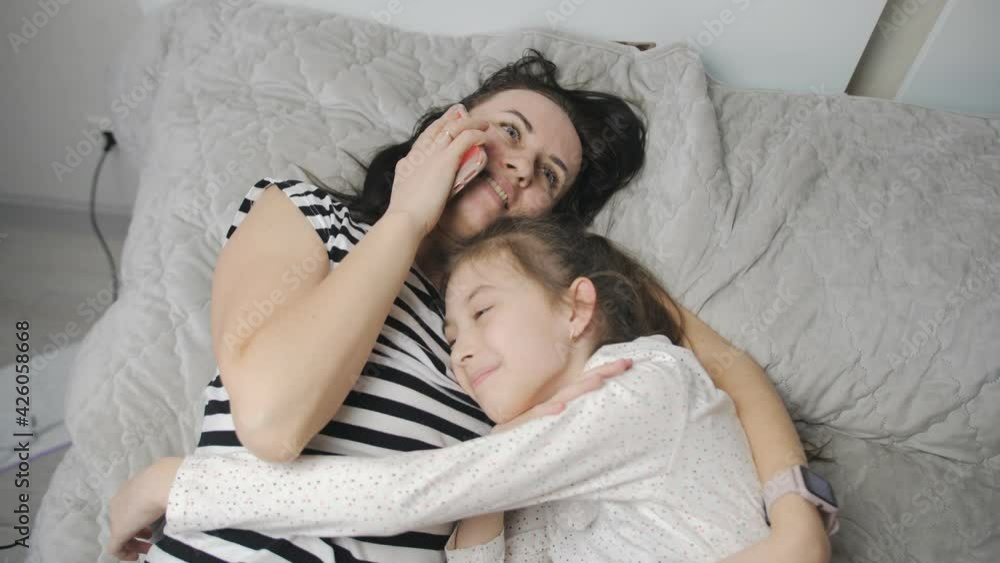 The daughter hugs her mother lying together on the bed and communicating on a mobile phone with relatives while in self-isolation. The daughter hugs her mother lying together on the bed and communicating on a mobile phone with relatives while in self-isolation.