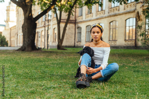 Attractive woman in casual clothes sitting on the grass in the park with a cute little dog in her arms and looking at the camera.
