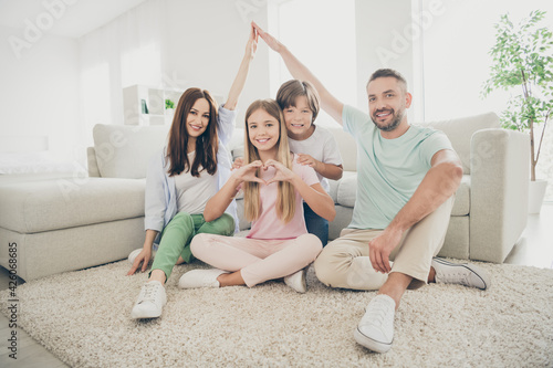 Photo portrait of parents showing roof with hands over heads and children sitting on carpet little daughter showing heart sign