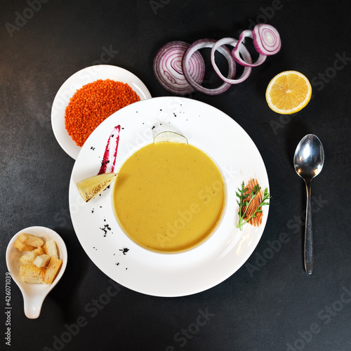 Plate of lentil cream soup with croutons on black table with ingredients for its preparation, Red lentils, cutting onion and lemon. Delicious and healthy lunch. Vegetarian diet food. High angle view.