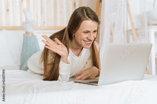 Freelance woman having a video call chat, typing at laptop and online shopping, lying on the white bed. Happy relaxed girl woking from home office. Distance learning online education and work.