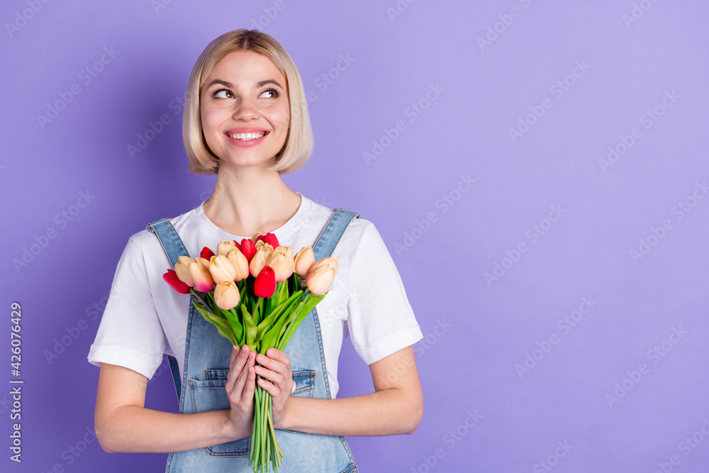 Photo of nice optimistic short hairdo blond lady hold flowers look empty space wear white t-shirt isolated on violet background Photo of nice optimistic short hairdo blond lady hold flowers look empty space wear white t-shirt isolated on violet background